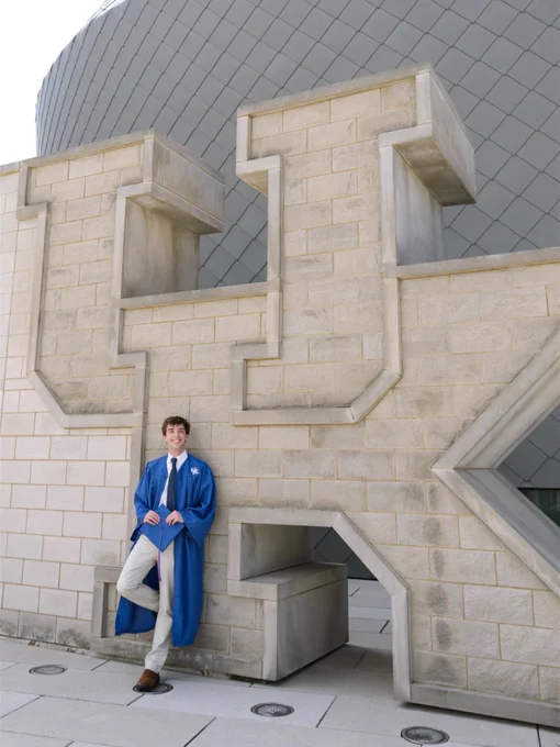 A person in a blue cap and gown is leaning against a large stone wall shaped like the letters "UK." The background features a modern building with a curved, metallic design.