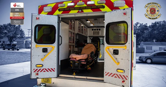 Open ambulance rear showing stretcher and medical equipment inside, with a suburban street in the background. Polk County Fire Rescue Logo in the top right. Text on American Heart Association Logo (Located on the top left): "American Heart Association. EMS. 2026 Gold. Target: Stroke Honor Roll. Target: Heart Attack Honor Roll. Mission: Lifeline.
