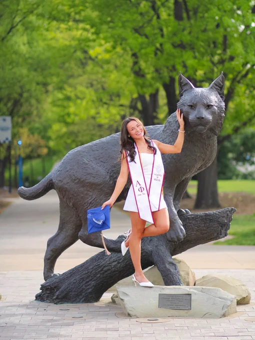 A person in a graduation outfit poses happily next to a large statue of the University of Kentucky's wildcat mascot. The scene is set outdoors with lush green trees in the background. The person holds a blue graduation cap.
