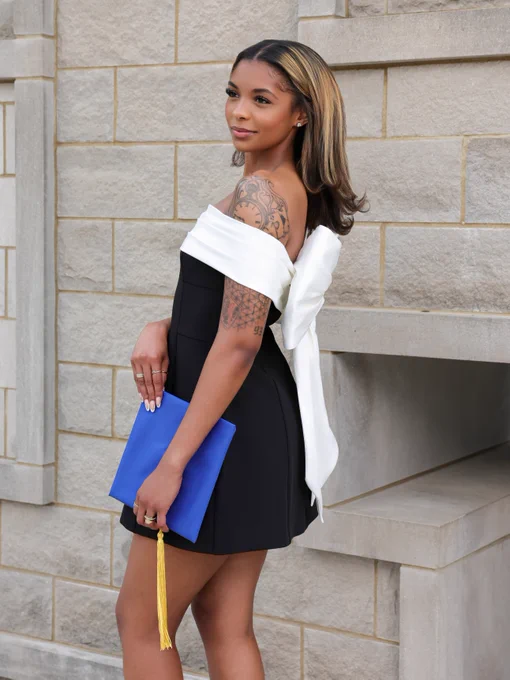 A person is dressed in a black and white dress and holds a blue graduation cap with a yellow tassel. They have tattoos on their arm and stand against a beige stone wall, looking to the side.