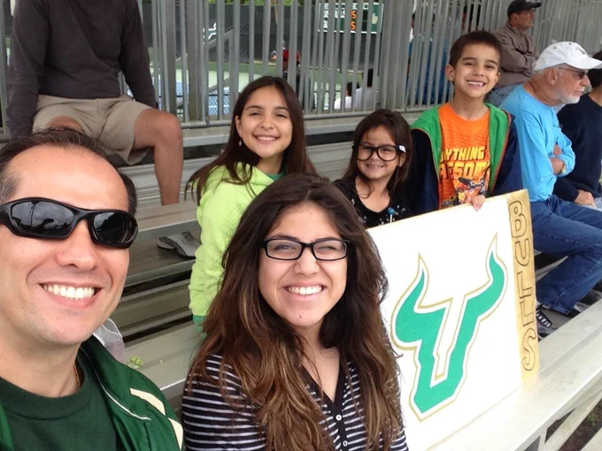 A group of people smiles at the camera while sitting in bleachers. One holds a sign with the USF Bulls logo. Some are wearing sunglasses and casual clothing. Others are in the background, with one wearing a baseball cap. Sara Arroyave Soto is in the back left.