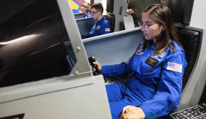 Sara Arroyave Soto in a blue NASA flight suits operates a simulator. American flag patches are visible on the suits.