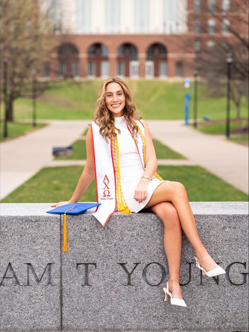 Person in a white dress and graduation stoles sits in front of William T. Young Library with a blue graduation cap beside them.