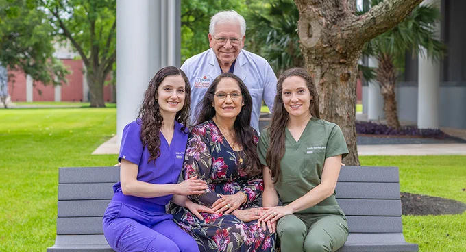 Three healthcare professionals, two women in scrubs and one man in a white coat, pose outdoors on a bench with a woman in a floral dress seated in front of them.