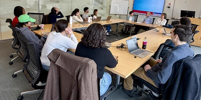 A group of people sit around a large conference table with laptops and notebooks. All are listening. There is a whiteboard in the background. The room has glass walls.