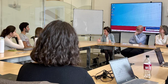 A group of people are seated around a conference table in a meeting room. One person is speaking while others listen attentively. A blank whiteboard and a large screen displaying a Microsoft Windows desktop are visible in the background. Several laptops and a bottle of Coca-Cola are on the table.