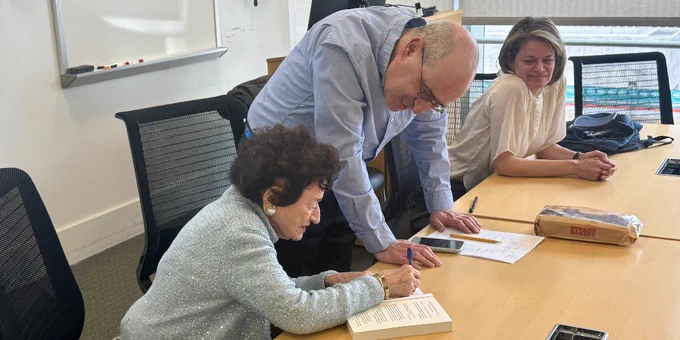 A person is sitting at a table signing a book while another person leans over to assist. A third person sits nearby, smiling. There's a whiteboard in the background and a bag on the table.