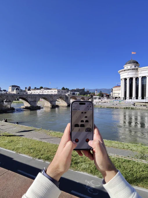 Hands holding an iPhone displaying RetroRapid mid-game, with a score of 19 visible on screen. In the background, the Stone Bridge and the Archaeological Museum of Macedonia are seen along the Vardar River in Skopje, under a clear blue sky with the North Macedonian flag visible on the museum dome.