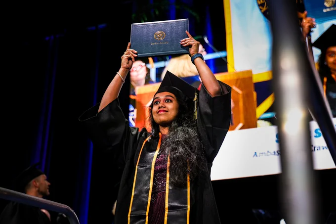 A Kent State graduate holds her diploma above her head with both hands and smiles on stage during commencement, wearing black cap, gown and gold honor stole.