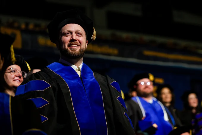 A person in academic regalia stands smiling during a graduation ceremony. The gown is black with blue accents. Other attendees in similar attire are in the background, with an indistinct stadium setting.