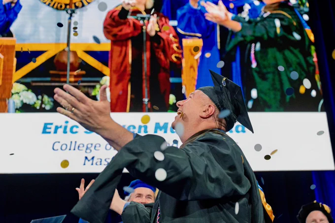 A person in a graduation cap and gown is joyfully celebrating while confetti falls. An out-of-focus background shows a large screen displaying a ceremony with additional graduates.