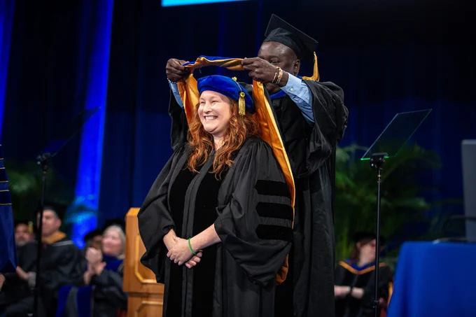 A graduate is being hooded during a commencement ceremony on stage. The setting includes academic regalia and an audience in the background. The atmosphere is formal and celebratory.