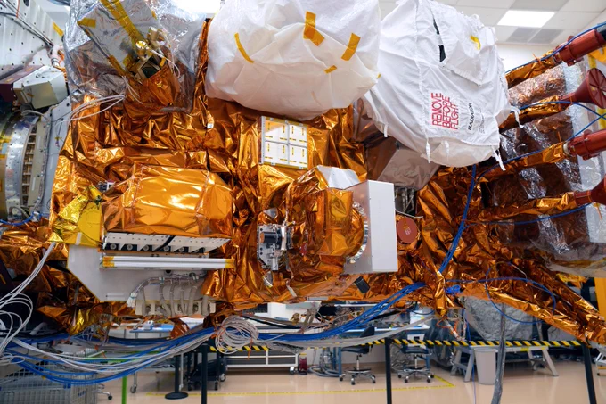 A view of a section of the JPSS-4 satellite, featuring the integrated Libera instrument. The spacecraft and its components are heavily covered in bright golden-orange thermal insulation. Several protruding parts are wrapped in white protective covers, one of which features bold red text reading \"Remove before flight.\" Bundles of blue and white cables hang loosely beneath the satellite body. The background shows a softly blurred cleanroom setting with work chairs, tables, and a black-and-yellow caution tape.
