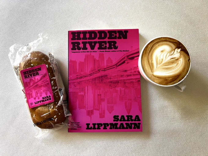HIDDEN RIVER beside the cutest book merch ever—a Philly soft pretzel with a Hidden River logo on the packaging. Beside a cappuccino