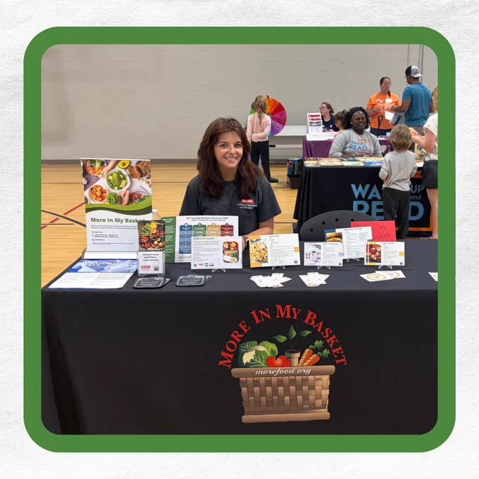 A woman sits at a table with "More In My Basket" branding, surrounded by pamphlets and cards.