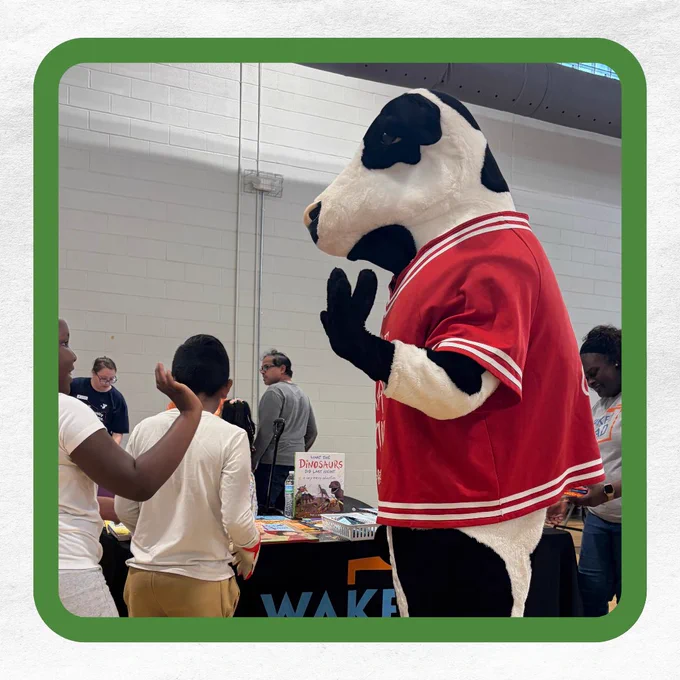 A person in a cow costume wearing a red jersey stands beside a table with books and people.