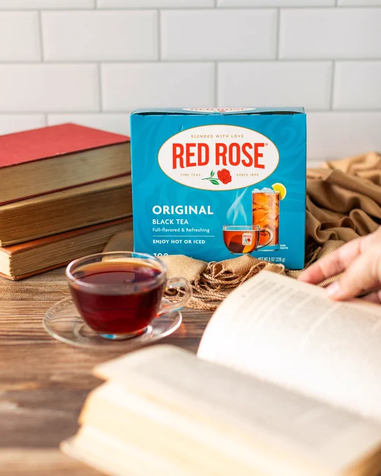 A cup of brewed black tea sits on a wooden table next to an open book and a box of Red Rose Original Black Tea.
