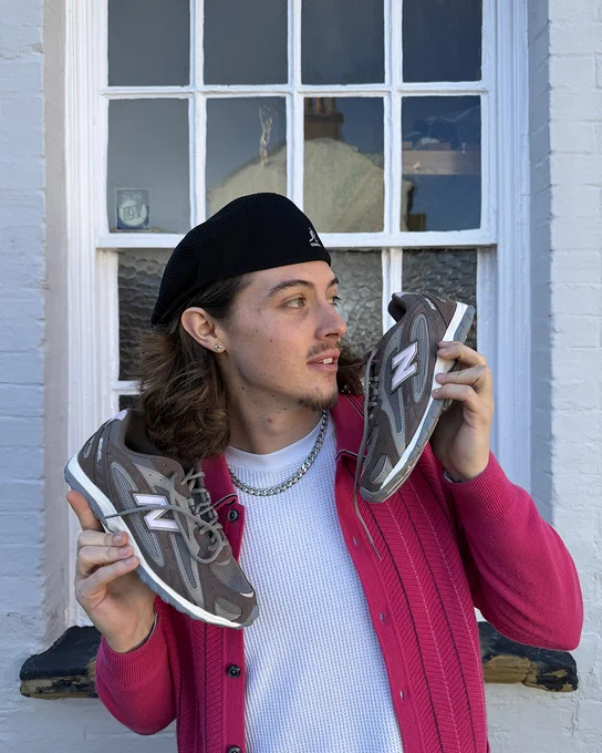 An image of a person holding a pair of brown New Balance 204L trainers in their hands whilst standing in front of a window.