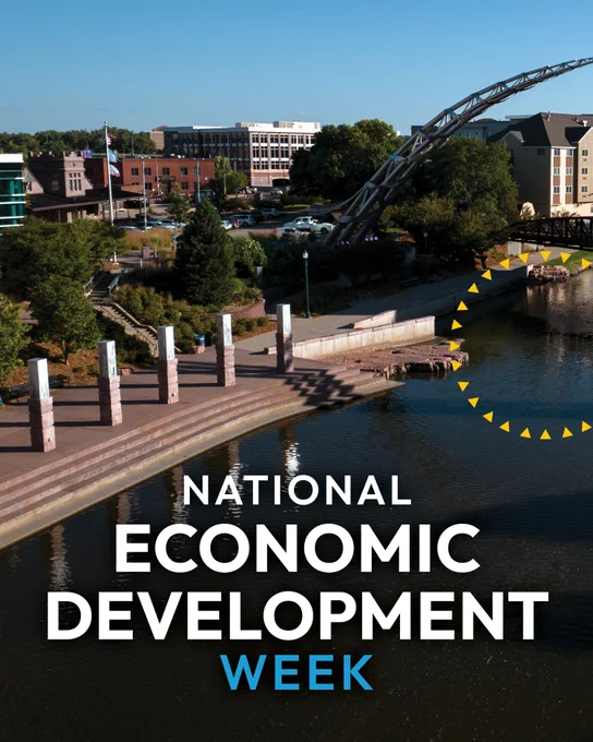 A scenic view of a riverside urban area featuring modern buildings and a unique arched sculpture. The water reflects the surroundings, and the text "National Economic Development Week" is displayed prominently in the foreground.