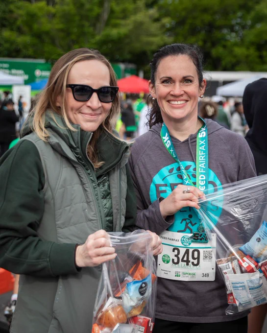 Two women at a charity event hold bags of donated food items. The mood is positive and community-focused.
