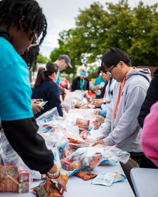 A group of volunteers gather outdoors around a table to assemble bags of food for distribution. The scene conveys teamwork and community service.