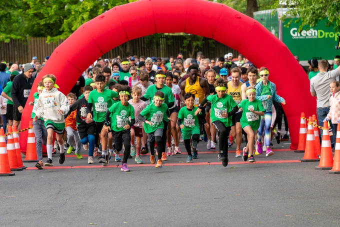 Excited children and adults gather at the starting line of a fun run race under a large red arch. The atmosphere is energetic and lively.
