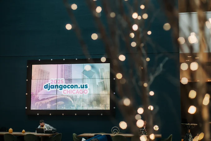 Screen displaying “DjangoCon US Chicago 2025” in a modern indoor space, with attendees seated at tables and decorative lights in the foreground.
