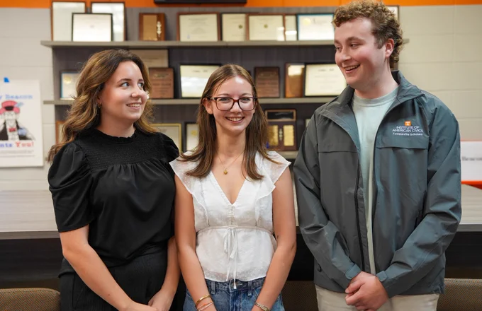 A group of three people pose in front of a wall of awards. The girl on the left has brown hair and is wearing a black top and black pants and is smiling at the two people next to her. The girl in the middle has brown hair and is wearing a white top with glasses and jeans. The guy on the far right has brown hair and is wearing a gray jacket and khaki pants.