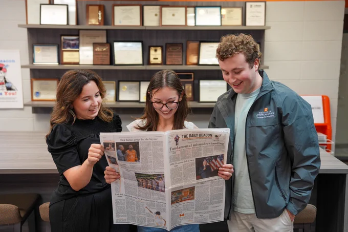 A group of three people standing in front of a wall of awards holding a newspaper looking at it. The girl on the left has brown hair and is wearing a black top and black pants and is smiling at the two people next to her. The girl in the middle has brown hair and is wearing a white top with glasses and jeans. The guy on the far right has brown hair and is wearing a gray jacket and khaki pants.