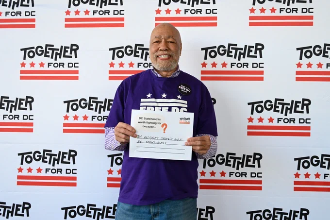 A Black man in a purple Free DC t-shirt smiles at the camera and holds a sign that says "DC statehood is worth fighting for because DC residents should not be second class."