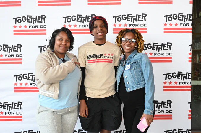 Three Black people stand in front of a backdrop with the Together for DC logo while smiling at the camera.