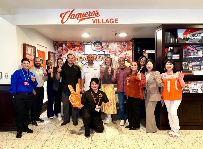 A group of about 12 people pose together inside a decorated “Vaqueros Village” lounge area featuring UTRGV sports memorabilia and orange-and-blue branding. Several people smile and hold up peace signs, while one person kneels in front holding a large orange foam hand. The space includes framed jerseys, banners, shelves with photos and décor, and a large UTRGV mural in the background.