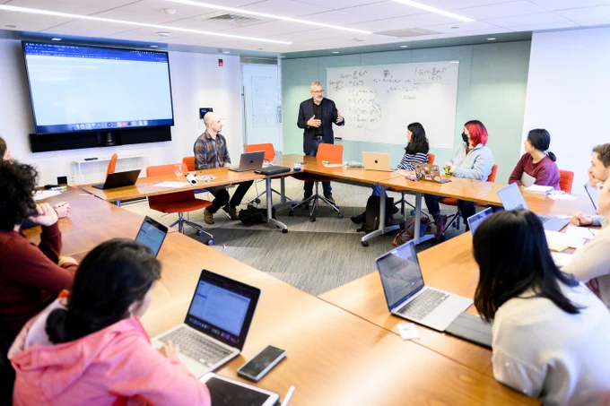 Office scene: Network Science professor gestures at a whiteboard covered in formulas while others sit around a U-shaped table with laptops. A large screen displays content at the front of the room. 
