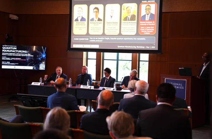 Patrick McCluskey sits on a panel with a screen reading "Quantum Manufacturing" behind him