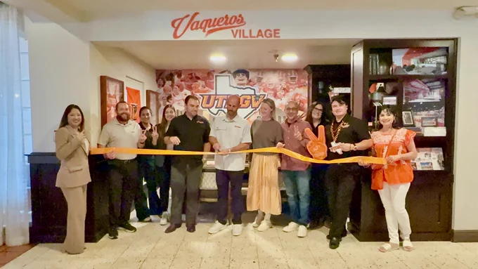 A group of people stand together for a ribbon-cutting ceremony inside the “Vaqueros Village” lounge area decorated with UTRGV athletics branding and memorabilia. Two people in the center cut a long orange ribbon while others smile, pose with peace signs, and hold a large orange foam hand. The background features a large UTRGV mural, framed jerseys, banners, and dark wood shelving with photos and sports décor.