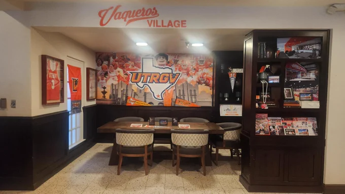 An empty “Vaqueros Village” seating area decorated with UTRGV athletics branding and memorabilia. The room features a large wall mural with the UTRGV logo and sports imagery, a wooden table surrounded by upholstered chairs, framed jerseys and banners on the walls, and dark wood shelving displaying books, photos, pennants, and magazines.
