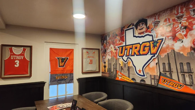 An empty “Vaqueros Village” seating area decorated with UTRGV athletics branding and memorabilia. The room features a large wall mural with the UTRGV logo and sports imagery, a wooden table surrounded by upholstered chairs, framed jerseys and banners on the walls, and dark wood shelving displaying books, photos, pennants, and magazines.