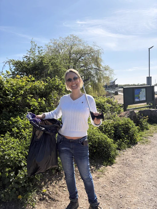A person standing outdoors, smiling and holding a bin bag and a picking tool. They are near greenery and a park information sign.