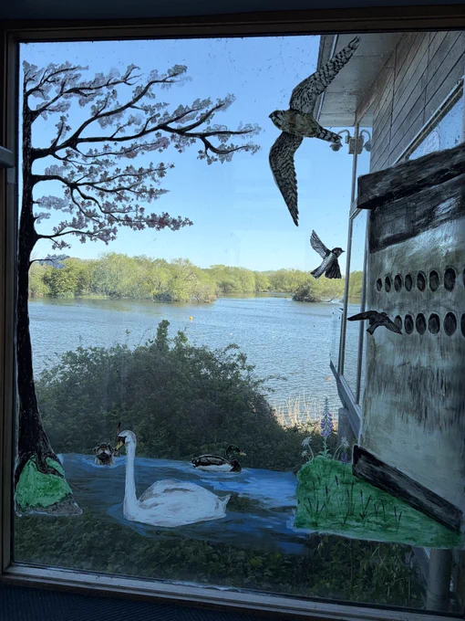 A window features paintings of birds, a tree, and a swan on a pond. Beyond, a scenic view of a lake and lush greenery is visible.