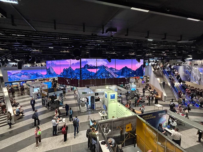 Wide view of a busy conference exhibition hall with research stands, visitors walking and talking, and a large multi-screen display showing a stylised mountain landscape overhead.