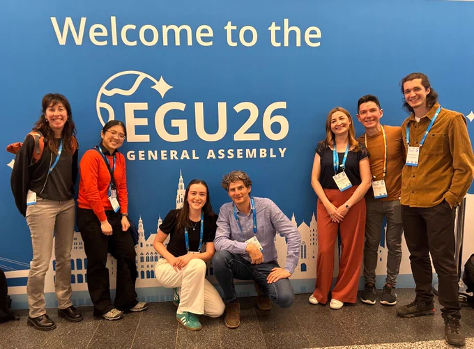 Group of conference delegates standing and kneeling in front of a large blue “EGU26 General Assembly” welcome backdrop, wearing name badges and lanyards.