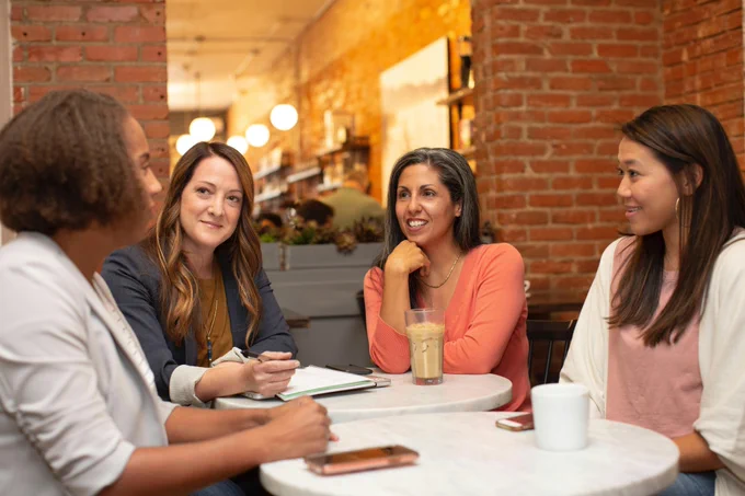 Four women of diverse backgrounds meeting at tables in a cafe, with drinks and notebooks.