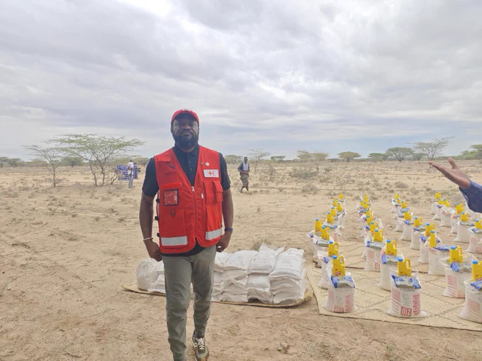 A person is standing in an arid landscape wearing a red vest with the International Federation of Red Cross and Red Crescent Societies logo. Nearby, there are arranged supplies, possibly for distribution. The sky is overcast.