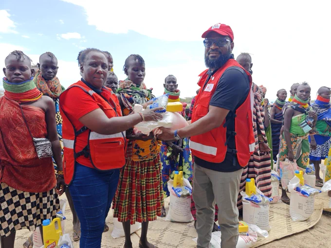 A group of people from the Turkana community stand outdoors in a line. Two individuals wearing Red Cross vests are distributing aid packages. Multiple containers and bags are placed on the ground. The sky is clear and blue in the background.