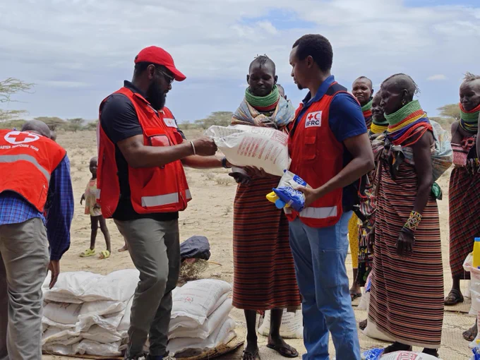 A group of people in a dry landscape are receiving aid supplies. Two individuals in red vests with the Red Cross logo are distributing bags and other items to locals wrapped in colorful garments. There are more bags stacked on the ground nearby.