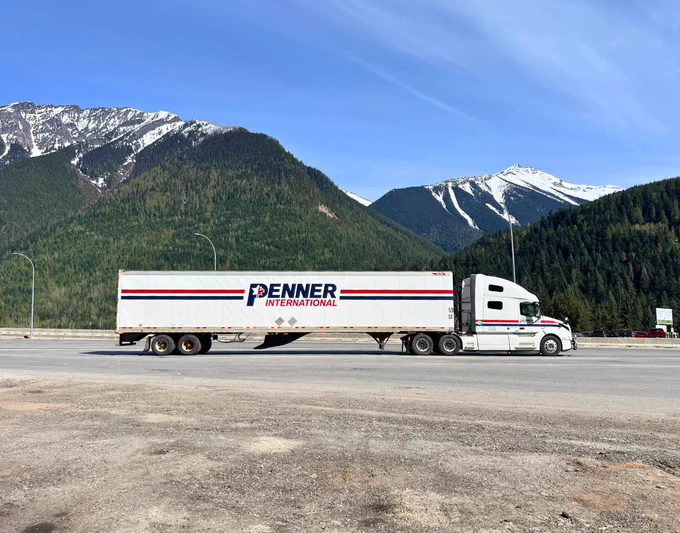 Penner International semi truck with a white trailer parked on a roadside pull-off, set against tall green forested mountains and snow-capped peaks under a clear blue sky.