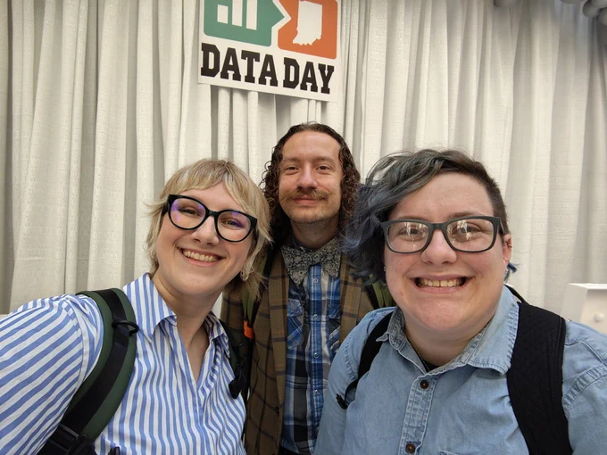 Three people are smiling at the camera in front of a sign that reads "Data Day." They are wearing glasses and casual clothing.