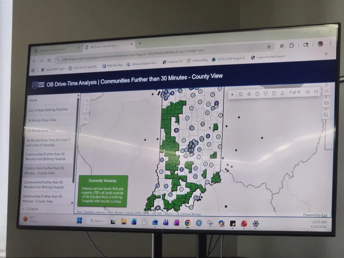 A large monitor displays a map labeled "OB Drive-Time Analysis | Communities Further than 30 Minutes - County View." The map shows various regions with symbols and color‑coded areas, primarily in green. A pop‑up on the map provides additional data. Several icons are positioned at the bottom of the screen.