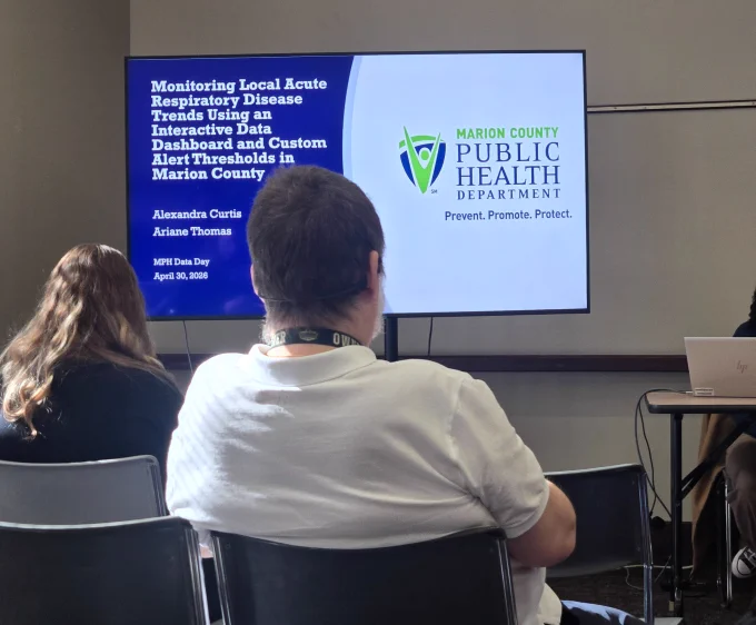 A presentation is taking place in a room with several attendees. A large screen displays a slide titled "Monitoring Local Acute Respiratory Disease Trends Using an Interactive Data Dashboard and Custom Alert Thresholds in Marion County." The slide also shows the Marion County Public Health Department logo and the date, April 30, 2020, with the names Alexandra Curtis and Ariane Thomas.