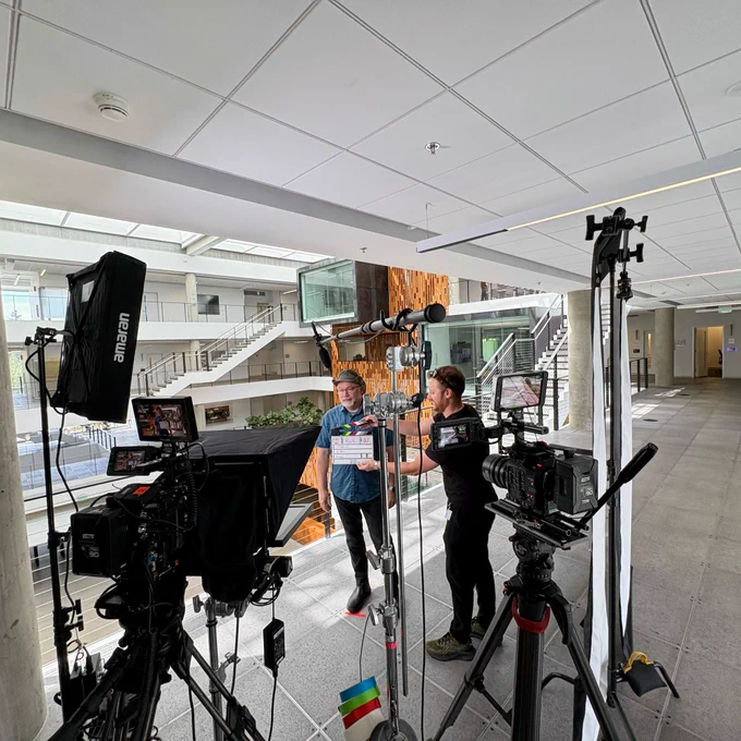 Rick Claus wearing his green hat standing  on a mobile video record set. Multiple cameras, multiple lights, and microphones are strewn around the scene.

The stage overlooks the atrium in a modern building. A sound guy with a timecode clacker is in front of Rick's chest.